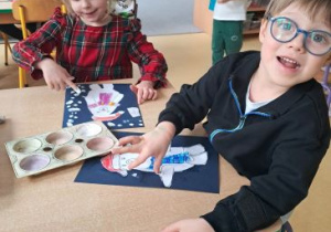 Dzieci siedzą przy stoliku i przyklejają wycięte elementy