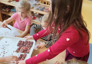 Dzieci siedzą przy stolikach, dziewczynka podchodzi do nich i częstuje zrobioną przez siebie walentynkową czekoladą