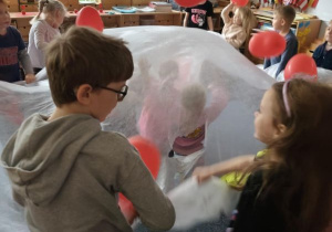 Dzieci trzymają w dłoniach biały materiał podrzucając leżące na nim czerwone balony w kształcie serduszek