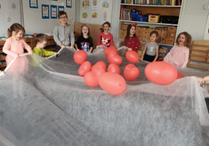 Dzieci trzymają w dłoniach biały materiał podrzucając leżące na nim czerwone balony w kształcie serduszek