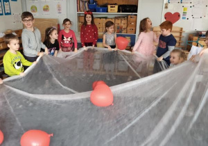 Dzieci trzymają w dłoniach biały materiał podrzucając leżące na nim czerwone balony w kształcie serduszek