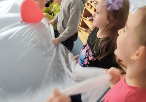 Dzieci trzymają w dłoniach biały materiał podrzucając leżące na nim czerwone balony w kształcie serduszek