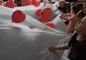 Dzieci trzymają w dłoniach biały materiał podrzucając leżące na nim czerwone balony w kształcie serduszek