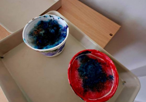 Zdjęcie przedstawia tace z kubeczkami wypełnionymi pomalowanym śniegiem przez dzieci w czasie zabawy badawczej