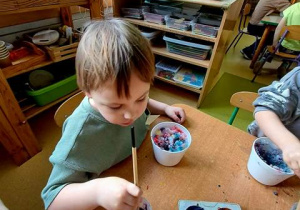 Chłopiec w czasie zabawy badawczej zamacza pędzel w kubeczku z zabarwioną farbami wodą