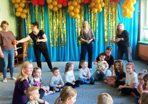 Dzieci siedzą na dywanie w kole a rodzice stoją dookoła nich