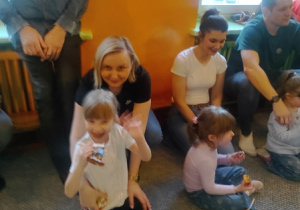 Rodzice siedzą z dziećmi na dywanie.