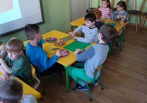 Dzieci siedzą przy stolikach, przed nimi leżą deski do krojenia, jajka, rzodkiewka i szczypiorek
