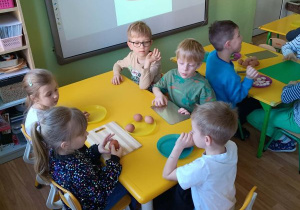 Dzieci siedzą przy stolikach, przed nimi leżą deski do krojenia i jajka