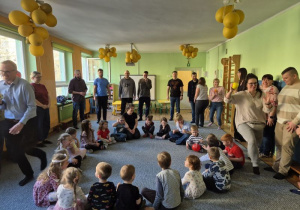 Dzieci siedzą w kręgu i podają sobie piłkę, rodzice stoją w większym kręgu dookoła dzieci i biegają z piłkami
