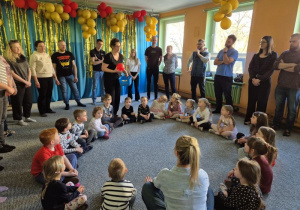 Dzieci siedzą w kręgu i podają sobie piłkę, rodzice stoją w większym kręgu dookoła dzieci i biegają z piłkami