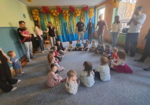 Dzieci siedzą w kręgu na dywanie, rodzice tworzą koło