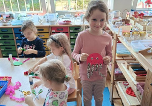Dzieci wykonują pracę plastyczną przy stolikach, obok stolika stoi dziewczynka i prezentuhe wykonana przez siebie meduzę