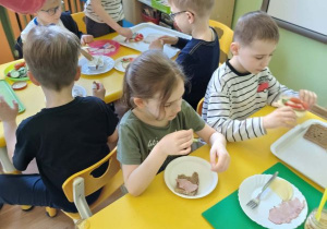 Dzieci siedzą przy stolikach i jedzą przygotowane przez siebie kanapki