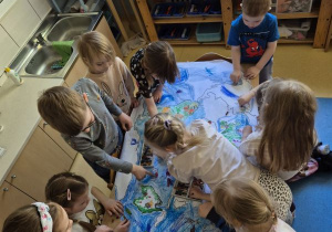 Dzieci stoją dookoła stołu i rysują wspólnie w grupie kilkuosobowej na dużym formacie