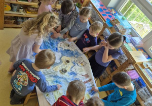 Dzieci stoją dookoła stołu i rysują wspólnie w grupie kilkuosobowej na dużym formacie