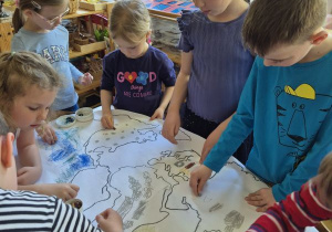 Dzieci stoją dookoła stołu i rysują wspólnie w grupie kilkuosobowej na dużym formacie
