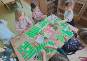 Dzieci siedzą przy stole i przyklejają kolejne elementy tulipanów