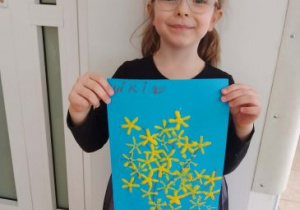 Dziewczynka pozuje do zdjęcia trzymając swoją pracę plastyczną przedstawiającą forsycję