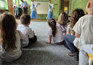 Dzieci siedzą i oglądają dwie tancerki ubrane w niebieskie sukienki podczas tańca