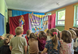 Dzieci siedzą na widowni i oglądają przedstawienie teatralne, na scenie stoi aktorka