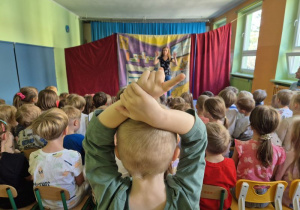 Dzieci siedzą na widowni i oglądają przedstawienie teatralne, na scenie stoi aktorka