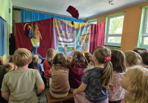 Dzieci siedzą na widowni i oglądają przedstawienie teatralne, na scenie stoi aktorka