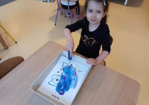 Dziewczynka siedzi przy stoliku i maluje tło do pracy plastycznej przy użyciu wałka