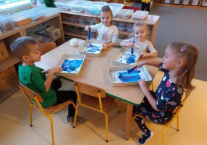 Dzieci siedzą przy stoliku i malują farbami tło do pracy plastycznej