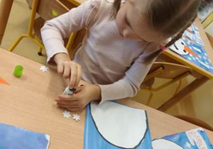 Dziewczynka siedzi przy stoliku i przykleja do przygotowanego tła kolorowe elementy kompozycji