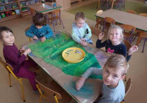 Grupa dzieci siedzi przy stoliku i koloruje tło plakatu z wykorzystaniem kredek pastelowych w różnych odcieniach koloru niebieskiego