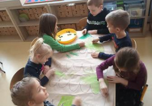 Grupa dzieci koloruje poszczególne elementy plakatu przedstawiającego choinkę