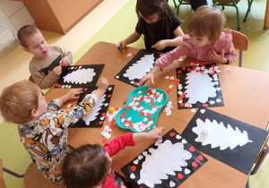 Dzieci siedzą przy stoliku i wykonują pracę plastyczną z wykorzystaniem gotowych elementów w kolorze biało-czerwonym