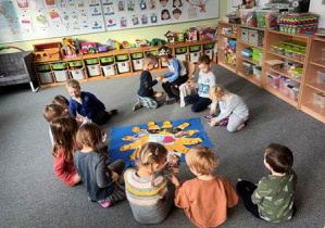 Dzieci siedzą na dywanie i oglądają zdjęcia Rysia z różnych etapów życia, w środku kręgu leży pomoc edukacyjna "Słoneczko" wykorzystywana w czasie urodzin dzieci