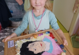 Dziewczynka trzyma korkową podkładkę z wykonanym przez siebie portretem Marii Montessori
