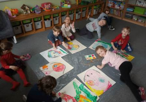 Dzieci siedzą na dywanie i malują farbami szkice narysowanych przez siebie portretów zainspirowanych twórczością Picasso