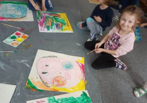 Dzieci na dywanie wypełniają tła swoich obrazów przy użyciu różnego koloru farb