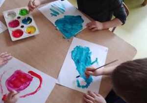 Dzieci młodsze siedzą przy stoliku i malują farbami ilustracje do słuchanego utworu A. Vivaldiego