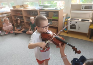 Chłopiec trzyma w dłoniach smyczek oraz skrzypce i próbuje zagrać na instrumencie